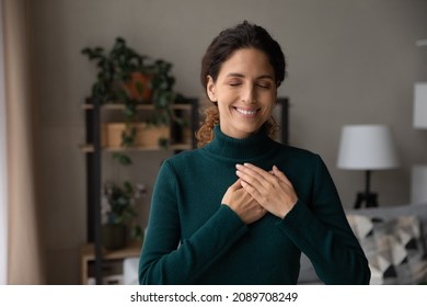Smiling young Hispanic woman hold hands at heart chest feel grateful and thankful. Happy millennial Latino female believer show love and care support pray or mediate. Faith, superstition concept.