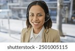 Smiling young hispanic woman wearing a headset in a professional setting with a blurred background.