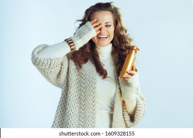 Smiling Young Housewife With Long Wavy Hair In Neck Sweater And Cardigan Holding Self Tanning Lotion Isolated On Winter Light Blue.