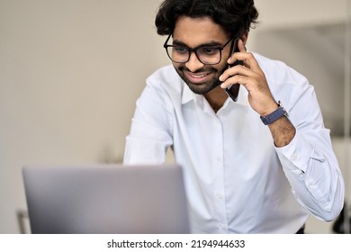 Smiling Young Indian Business Man Client Manager, Salesman Talking On Phone Consulting On Cellphone Making Call In Office Working Looking At Laptop Computer Making Bank Loan Offer, Selling Insurance