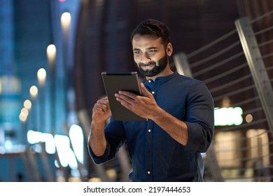 Smiling Young Indian Business Man Professional, Eastern Businessman Executive Standing Outdoors On Street Reading Ebook Holding Using Digital Tablet Online Technology In Night City With Urban Lights.