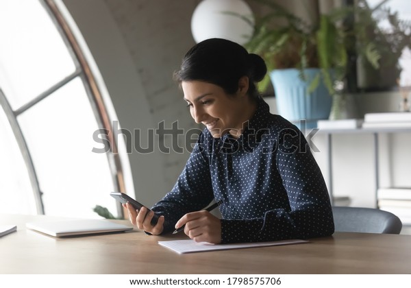 Smiling young indian businesswoman filling paper\
document form, web surfing important information online on\
smartphone. Distracted from paperwork happy mixed race employee\
chatting in social\
network.