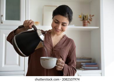 Smiling young indian ethnicity woman pouring hot water from kettle into cup, brewing tea or coffee, drinking beverage in modern kitchen, enjoying peaceful morning evening stress free carefree time. - Powered by Shutterstock