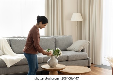Smiling young indian ethnicity woman homeowner putting beautiful flowers in vase on coffee table in modern living room, enjoying decorating own apartment, improving styling interior on weekend.