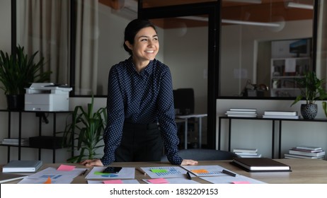 Smiling young indian female employee stand at desk in office look in distance thinking or visualizing career success. Happy ethnic businesswoman plan or dream at workplace. Business vision concept.