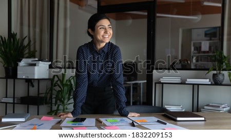 Smiling young indian female employee stand at desk in office look in distance thinking or visualizing career success. Happy ethnic businesswoman plan or dream at workplace. Business vision concept. Foto stock © 