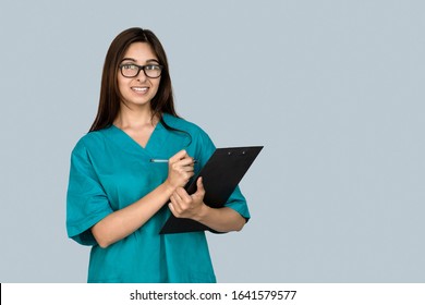 Smiling Young Indian Female Nurse Student Hospital Medical Staff Worker Wear Green Coat Write In Clipboard Prescribing Fill Information At Training Look At Camera Isolated On Gray Studio Background.