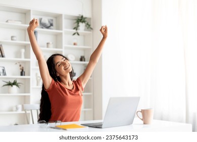 Smiling young indian lady freelancer stretching during working day at home office, millennial eastern woman sitting at desk with laptop computer, resting after finishing online project, copy space - Powered by Shutterstock