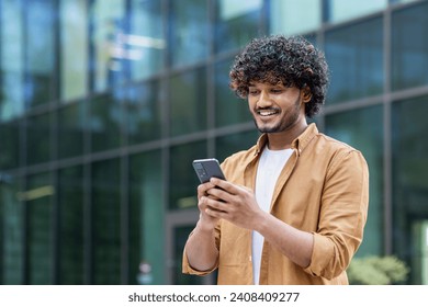 A smiling young Indian man is standing on the street near an office center and using the phone, talking on a video call. - Powered by Shutterstock