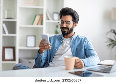 Smiling Young Indian Man Using Smartphone And Drinking Coffee At Home, Happy Millennial Eastern Guy Browsing Social Networks On Mobile Phone While Relaxing At Desk Indoors, Copy Space