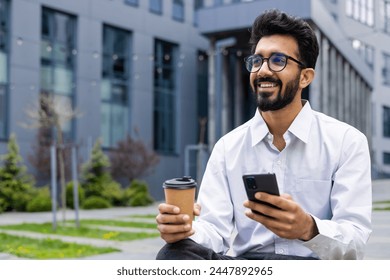 A smiling young Indian man in a white shirt and glasses is sitting outside a building, resting on a work break, holding a phone and a cup of coffee, and looking happily to the side. - Powered by Shutterstock