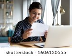 Smiling young indian woman holding paper bill letter doing paperwork bill reading good news check post mail sit at home table, happy lady customer receive bank receipt sheet tax refund notification