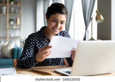 Smiling young indian woman holding paper bill letter doing paperwork bill reading good news check post mail sit at home table, happy lady customer receive bank receipt sheet tax refund notification