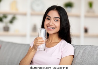 Smiling young Indian woman holding glass of fresh clean water while sitting on couch in living room. Lovely millennial lady taking care of her wellbeing and health. Keep hydrated concept