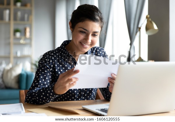 Smiling young indian woman holding paper
bill letter doing paperwork bill reading good news check post mail
sit at home table, happy lady customer receive bank receipt sheet
tax refund notification