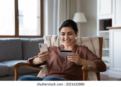 Smiling young Indian woman sit in chair at home shopping online on smartphone with credit debit card. Happy millennial ethnic female buyer make payment buy on internet use secure banking on cellphone.