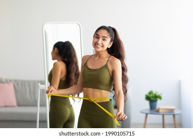 Smiling young Indian woman standing near mirror and measuring her waist at home. Potrait of pretty Asian lady feeling happy about her body measurements. Weight loss and dieting concept