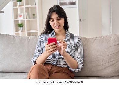 Smiling Young Indian Woman Using Smart Phone Sitting On Sofa At Home. Happy Lady Holding Cell Phone Doing Online Shopping, Ordering Delivery In Mobile App, Checking Social Media Or Dating Application.