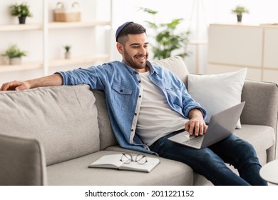 Smiling Young Jewish Man Relaxing On Comfortable Couch In Living Room Using Modern Laptop, Happy Millennial Male Resting On Cozy Sofa At Home, Watching Movie Or Video Online, Enjoying Weekend