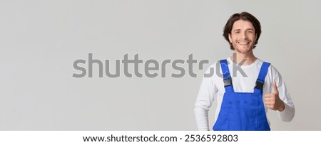 Smiling young laborer posing in work overalls and showing thumb up over light studio background, full-length shot, copy space