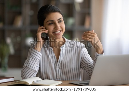 Smiling young lady working at home office desk by laptop engaged in phone conversation with client give remote consultation. Indian woman employee make business call from workplace distracted from pc