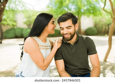 Smiling young latin couple sitting closely, whispering on ear and radiating happiness on their sunlit outdoor date - Powered by Shutterstock