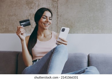 Smiling Young Latin Woman 20s In Casual Clothes Sit On Sofa Using Mobile Cell Phone Hold Credit Bank Card Doing Online Shopping Spend Time In Living Room Home In Own House Be Lost In Reverie Good Day