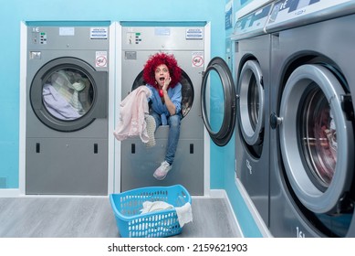 A Smiling Young Latin Woman With Afro Hair Sitting Inside A Washing Machine In A Blue Automatic Laundry Room