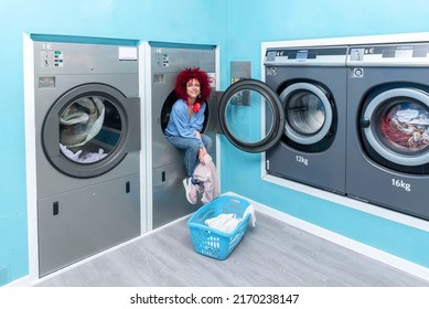A Smiling Young Latin Woman With Afro Hair Sitting Inside A Washing Machine In A Blue Automatic Laundry Room