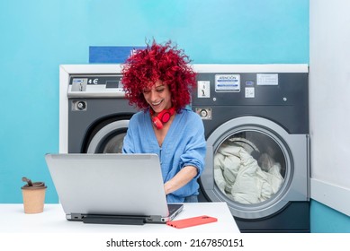 a smiling young latin woman with red afro hair works with her laptop in the blue laundry room while waiting for the laundry to be done, her red headphones around her neck,  - Powered by Shutterstock