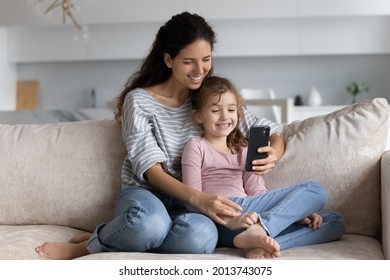 Smiling Young Latino Mom And Biracial Teen Daughter Rest On Couch At Home Have Webcam Virtual Event On Smartphone. Happy Hispanic Mother And Ethnic Girl Child Talk Speak On Video Call On Cellphone.