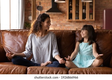 Smiling Young Latino Mother And Little Girl Child Sit On Sofa In Living Room Meditate Practice Yoga Together. Happy Hispanic Mom And Small Daughter Relax On Couch Exercise At Home Engaged In Activity.