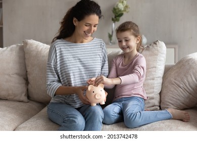 Smiling Young Latino Mother And Teen Daughter Sit On Sofa Drop Coin Cash Save Money In Piggybank. Happy Provident Economical Hispanic Mom And Small Girl Child Make Finance Investment For Future.