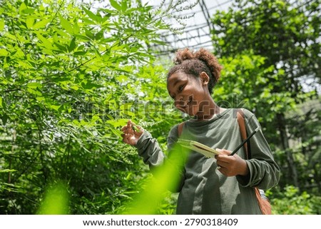 Smiling young learner exploring tropical vegetation inside a biodiversity dome while recording observations in a field notebook. This authentic eco education scene highlights scientific curiosity, environmental literacy, plant identification, ecosystem learning, and hands-on STEM education.