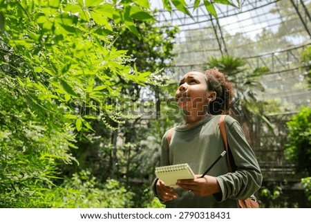 Smiling young learner exploring tropical vegetation inside a biodiversity dome while recording observations in a field notebook. This authentic eco education scene highlights scientific curiosity, environmental literacy, plant identification, ecosystem learning, and hands-on STEM education.