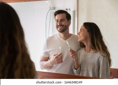 Smiling Young Loving Caucasian Couple Look In Mirror Enjoy Daily Morning Routine In Bathroom Together, Happy Millennial Newlyweds Brush Clean Teeth With Toothbrushes In Bath, Oral Hygiene Concept