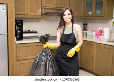 Smiling Young Maid Wearing Black Apron And Yellow Latex Rubber Gloves Standing In Kitchen Holding Black Plastic Garbage Bag