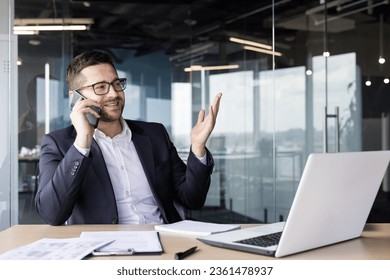 Smiling young male businessman talking on the phone with clients and partners sitting at the office desk in a suit. - Powered by Shutterstock