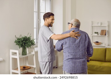 Smiling young male caregiver in uniform hugging elderly man during visit. Friendly social worker helping senior man with daily activities at home. Assistant, elderly people caregiver concept - Powered by Shutterstock