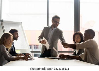 Smiling young male ceo executive manager giving out financial paper reports to diverse colleagues, analyzing sales data or developing company growth strategy at brainstorming meeting in office.