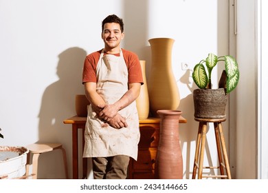 Smiling young male craftsman in apron leaning against edge of table with clay vases and looking at camera against lit background with shadows - Powered by Shutterstock