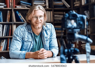 Smiling young male creator blogger influencer talking looking at professional dslr digital camera shoot education tutorial vlog training filming video course for social media sit at desk in library.