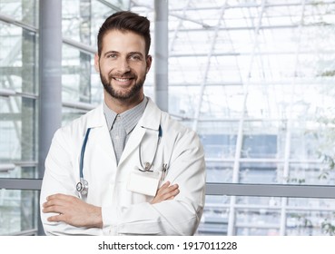 Smiling Young Male Doctor In Hospital, Looking At Camera Standing With Crossed Arms.