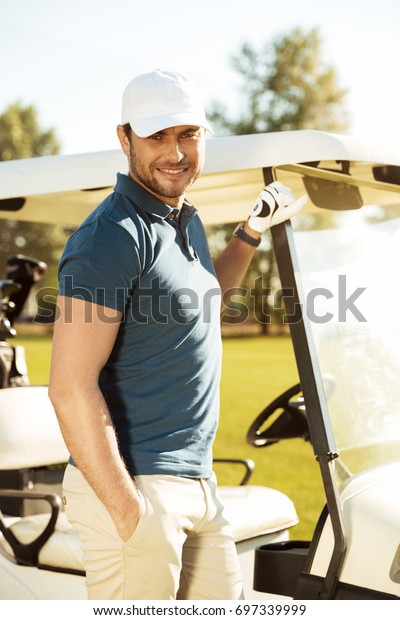 Smiling young male golfer standing at the golf\
cart and looking at\
camera