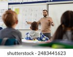 Smiling young male teacher helping students learn alphabet in first grader classroom. Rear view of children revising and learning letters in school classroom. Male teacher teaching alphabet to kids.