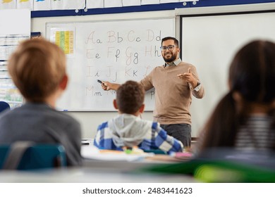 Smiling young male teacher helping students learn alphabet in first grader classroom. Rear view of children revising and learning letters in school classroom. Male teacher teaching alphabet to kids. - Powered by Shutterstock