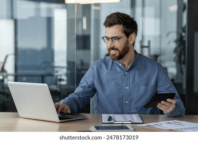 A smiling young man accountant, financial expert works in the office with documents, on a laptop, holding a calculator in his hands.