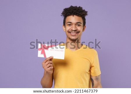 Smiling young man of African American ethnicity wear casual yellow t-shirt hold in hand gift coupon voucher card for store isolated on plain pastel light purple background studio. Mock up copy space