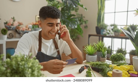 Smiling young man with apron holding credit card and talking on phone in flower shop. - Powered by Shutterstock