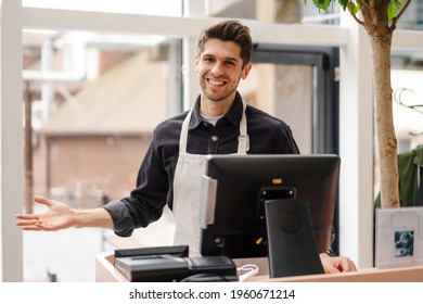 Smiling young man in apron standing at the cash register indoors - Shutterstock ID 1960671214